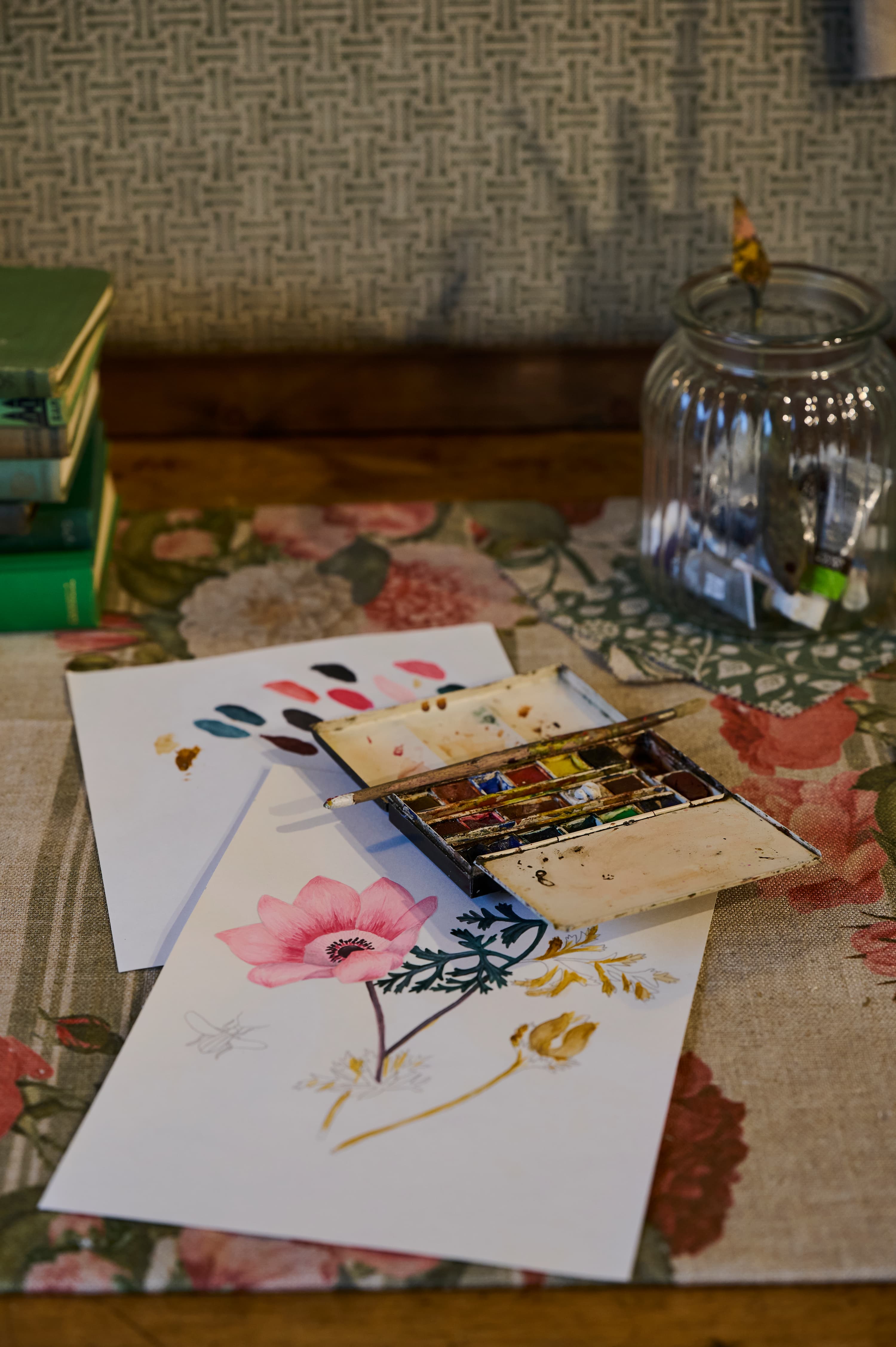 A watercolour painting of flowers on paper sits on a floral tablecloth, alongside a paint palette, colour swatches, a stack of green books, and a glass jar with a paintbrush in water.
